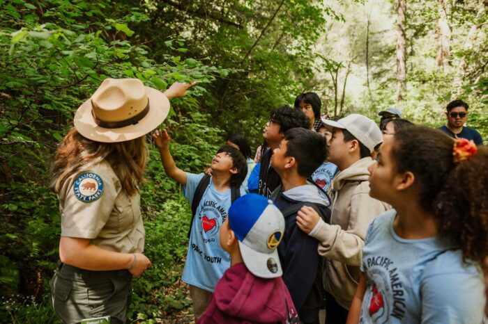 A group of school children look at and touch foliage along a trail with a state park ranger in the foreground.