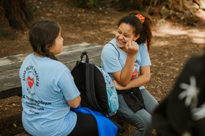 Two girls wearing Asisa Pacific American community Center T-shirts share a laugh while sitting on a bench in the forest