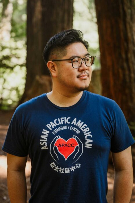 An Asian man wearing a blue Asian Pacific American Community Center T-shirt looks to his left, backed by redwood trees