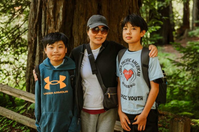 An Asian woman wearing sunglasses has her arms around the shoulders of her 12-year-old sons, one of whom wears an Asian Pacific American Community Center T-shirt.