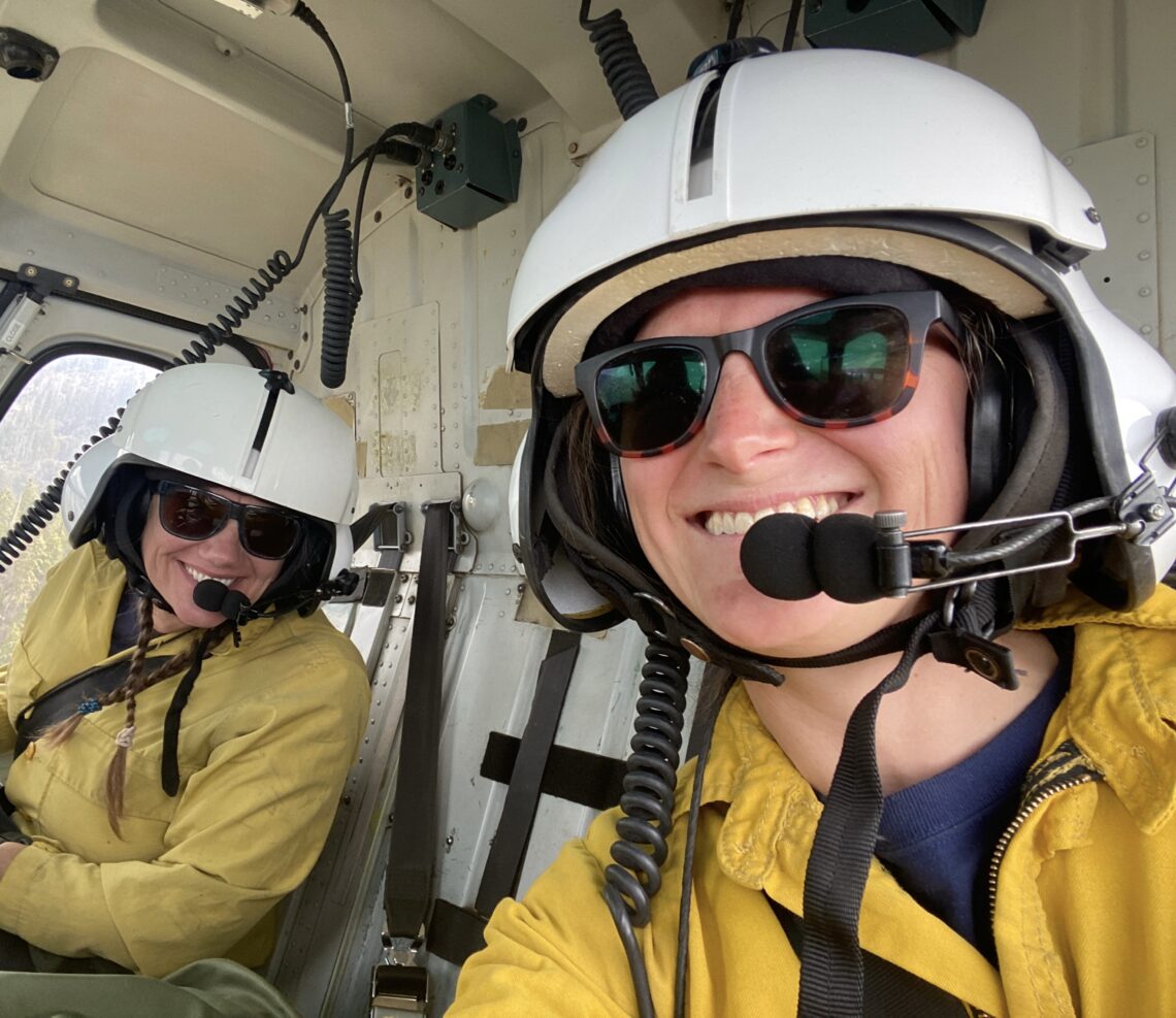 Two smiling women in a helicopter wearing white helmets and bright yellow safety jackets.