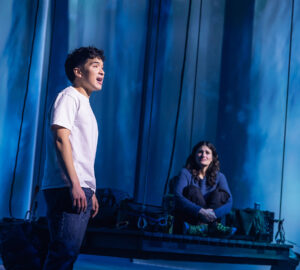 Zachary Noah Piser stands at the front of the stage in a white T-shirt, speaking or singing, while Idina Menzel sits on a platform in the background. The scene is illuminated with a cool blue forest backdrop.